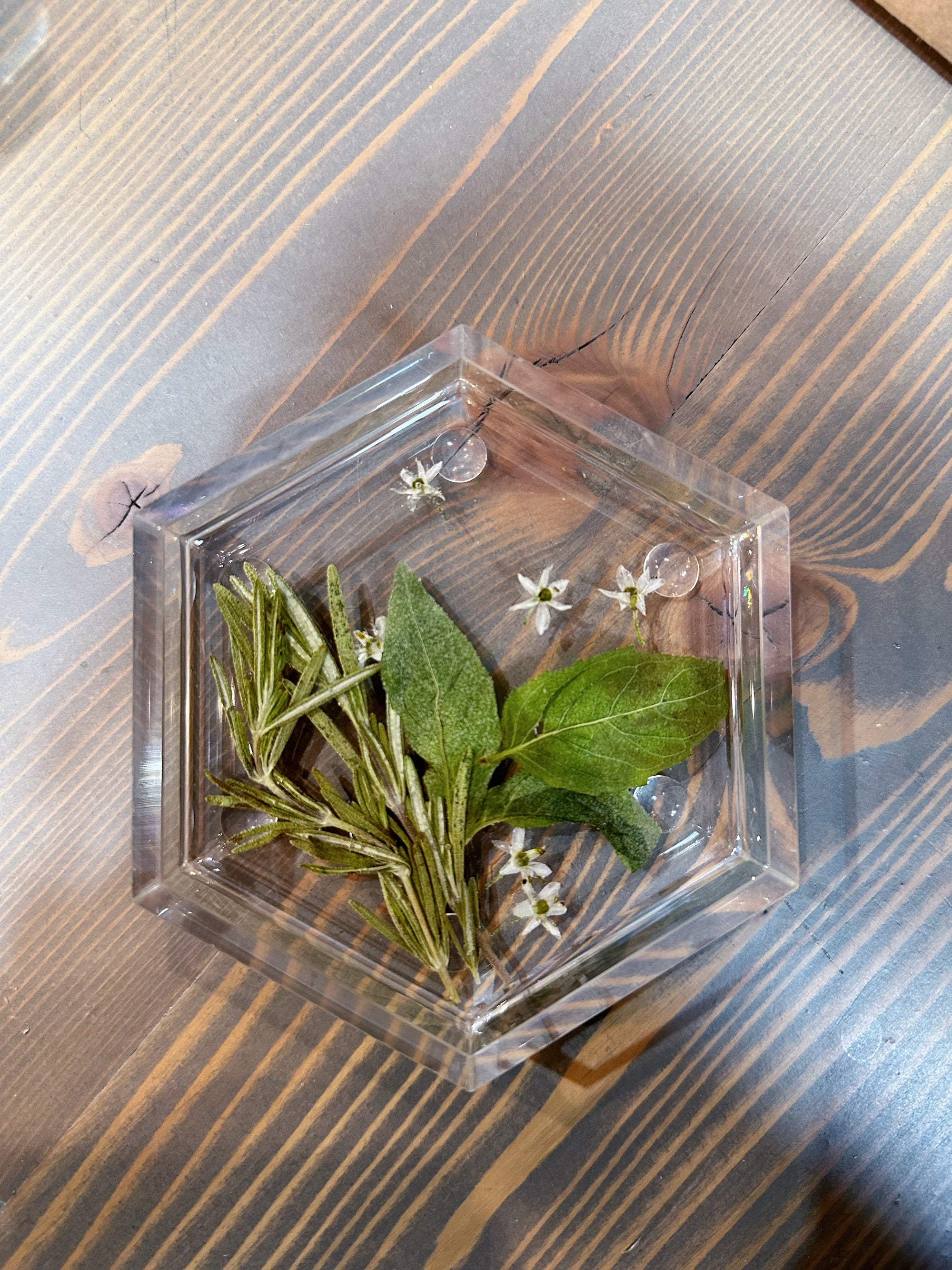 A hexagon resin ring dish features real pressed greenery and small white flowers. It is set against a wooden background. 