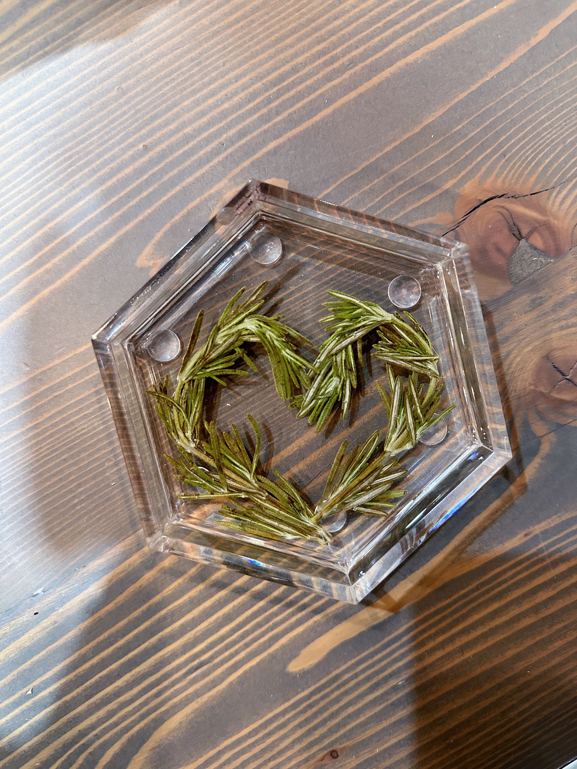 A resin hexagon ring dish featuring pressed greenery is designed in the shape of a heart and is set on top of a wooden surface.