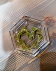 A resin hexagon ring dish featuring pressed greenery is designed in the shape of a heart and is set on top of a wooden surface.