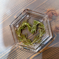 A resin hexagon ring dish featuring pressed greenery that is shaped like a heart, set on top of a wooden surface.