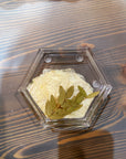 A hexagon resin ring dish with a pressed white flower and some greenery is placed on top of a wooden surface. 