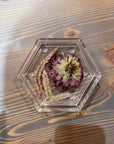 A hexagon resin ring dish with a purple pressed flower and a few pink flowering stems is set on top of a wooden surface. 