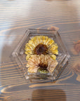 A hexagon resin ring dish with a pressed yellow and cream flowers is placed against a wooden background. 