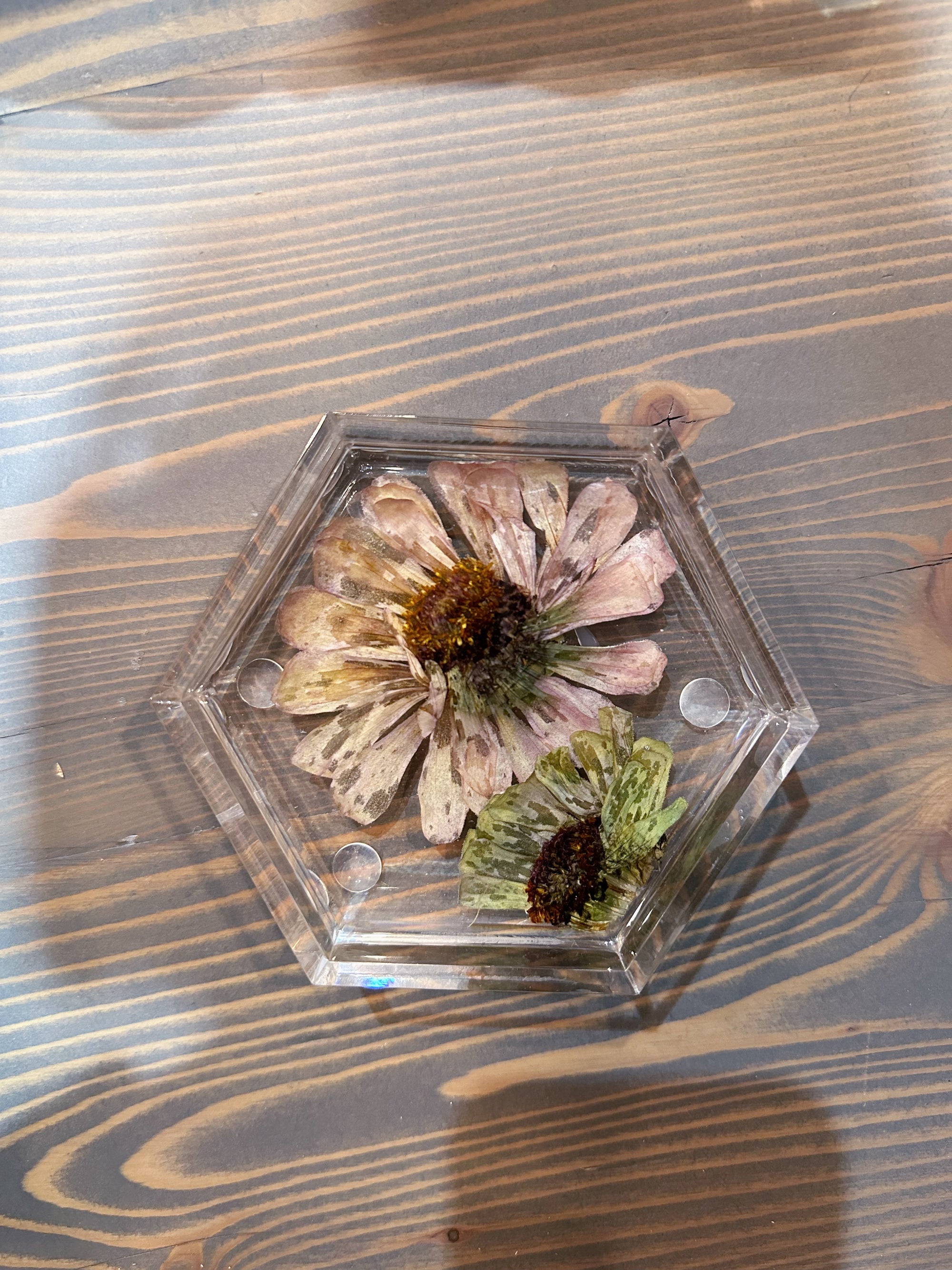 A hexagon resin ring dish with one pressed pink flower and one small green flower is set against a wooden background. 