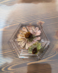 A hexagon resin ring dish with one pressed pink flower and one small green flower is set against a wooden background. 