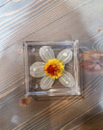 A square resin ring dish includes a pressed white flower with a yellow and orange flower placed at its center. It is set on a wooden surface. 