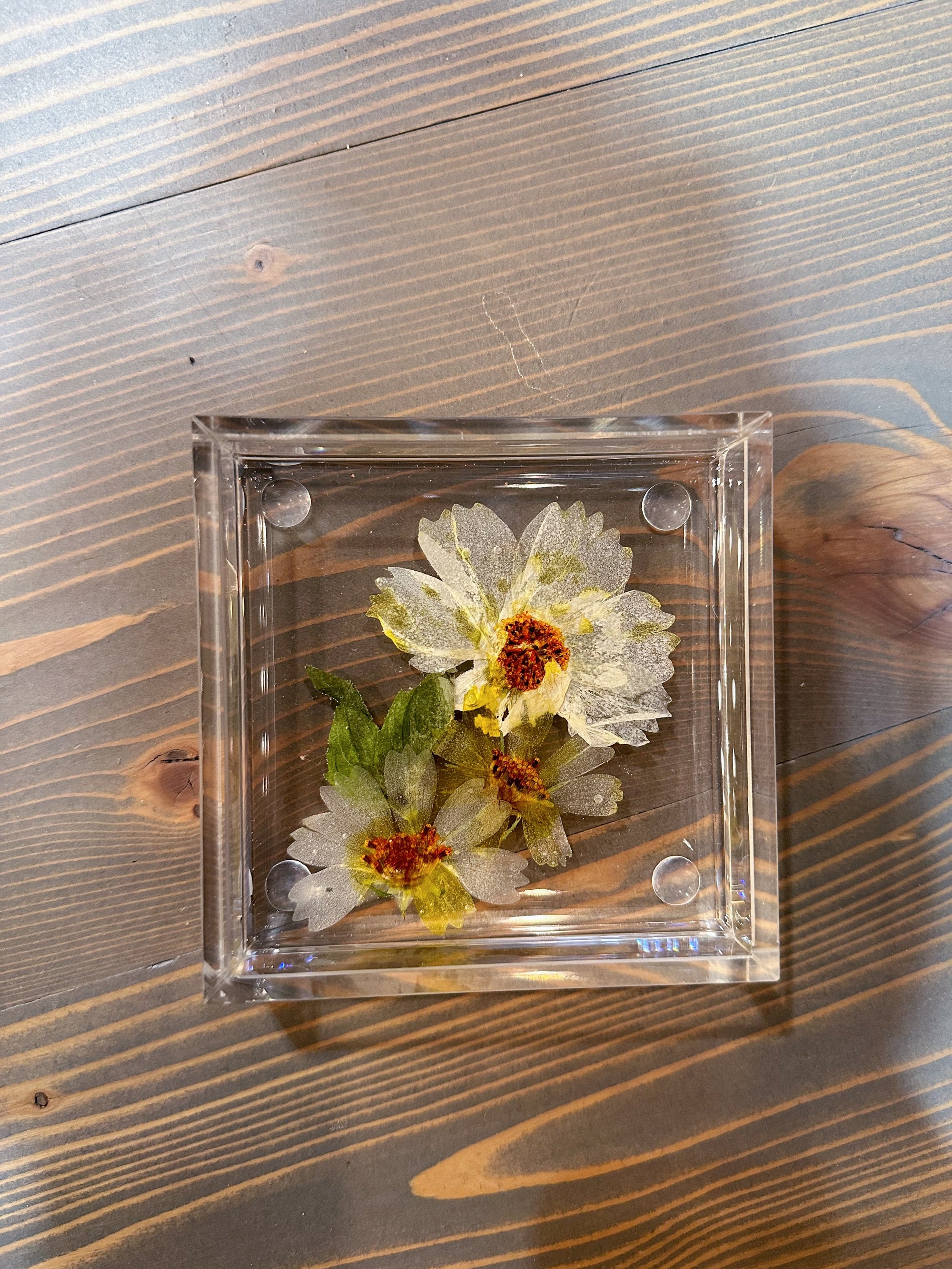 A square resin ring dish with small pressed white flowers is placed on top of a wooden surface. 