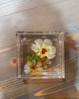 A square resin ring dish with small pressed white flowers is placed on top of a wooden surface. 