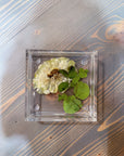 A square resin ring dish with one pressed white flower and greenery is set atop a wooden surface. 