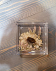 A square resin ring dish designed with one pressed white flower and a few flowering greens is set against a wooden background. 