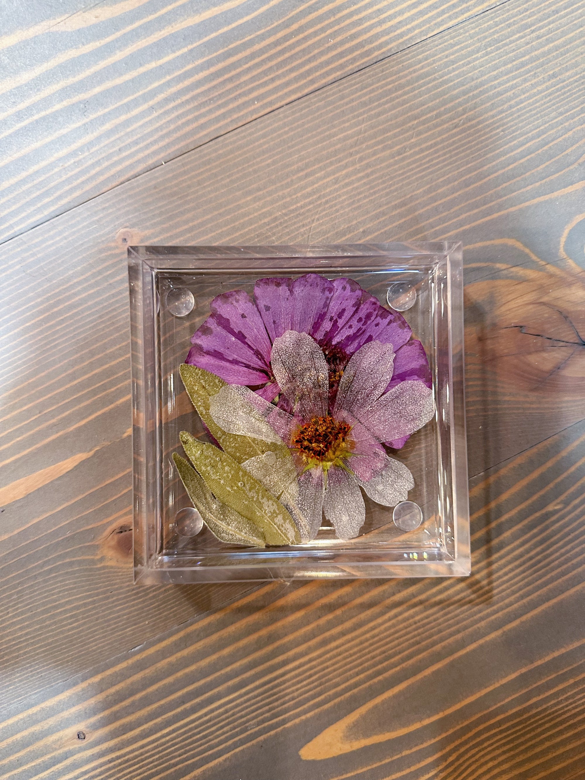 A square pressed flower resin ring dish features one purple flower placed behind a transparent white flower a few green leaves. It is set atop a wooden surface. 