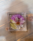 A square pressed flower resin ring dish features one purple flower placed behind a transparent white flower a few green leaves. It is set atop a wooden surface. 