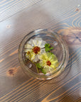 A circle resin ring dish with two pressed white and yellow flowers is placed on top of a wooden surface. 