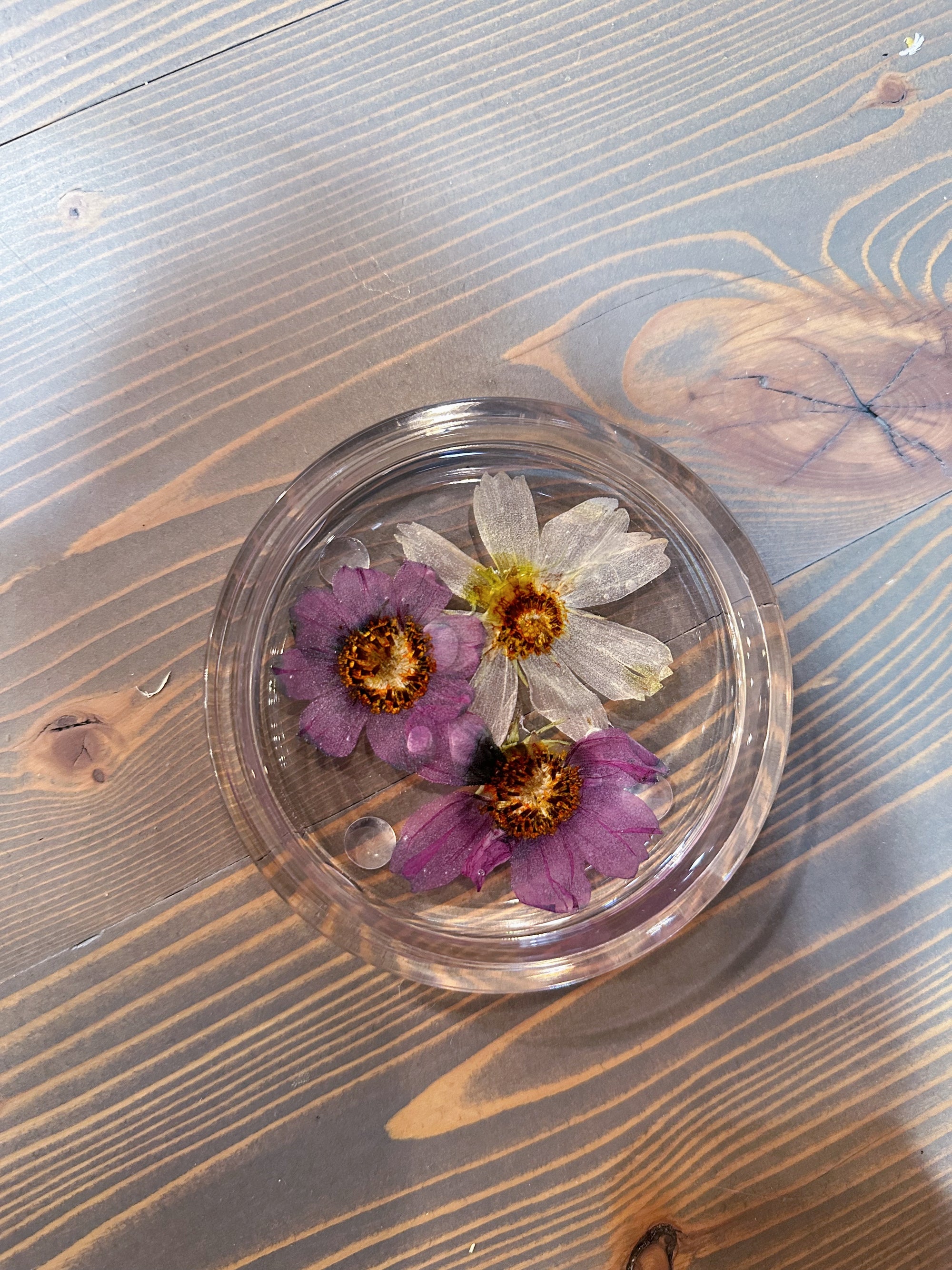 A circle resin ring dish designed with two pressed purple flower and one white flower is set against a wooden background. 