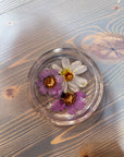 A circle resin ring dish designed with two pressed purple flower and one white flower is set against a wooden background. 