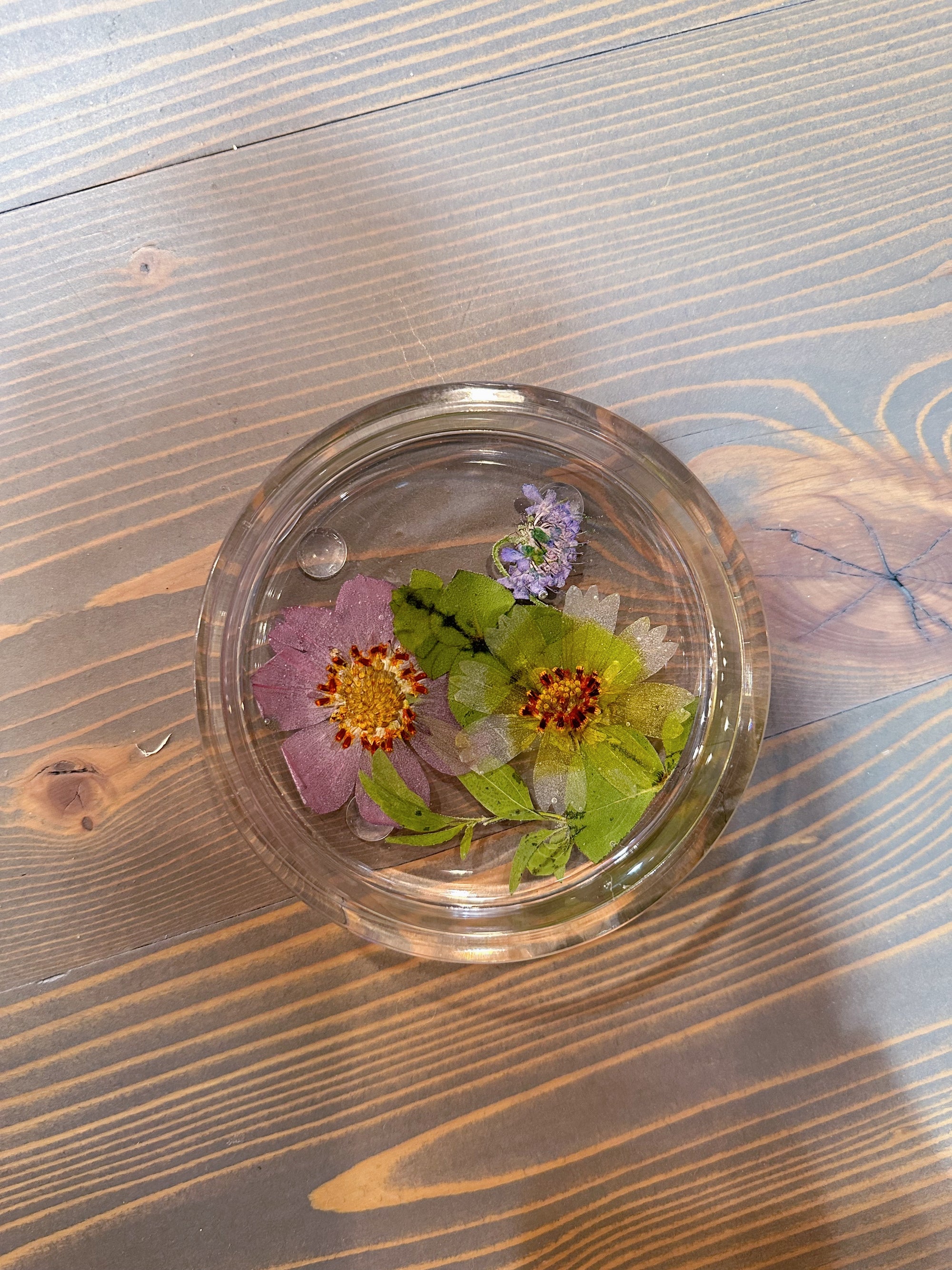 A circle resin ring dish includes pressed pink, green, and purple flowers and a few leaves. It is set against a wooden background. 