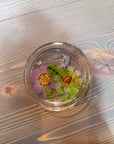 A circle resin ring dish includes pressed pink, green, and purple flowers and a few leaves. It is set against a wooden background. 