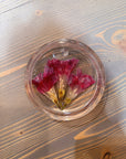 A circle resin ring dish with three pressed red and pink flowers is placed on top of a wooden surface. 