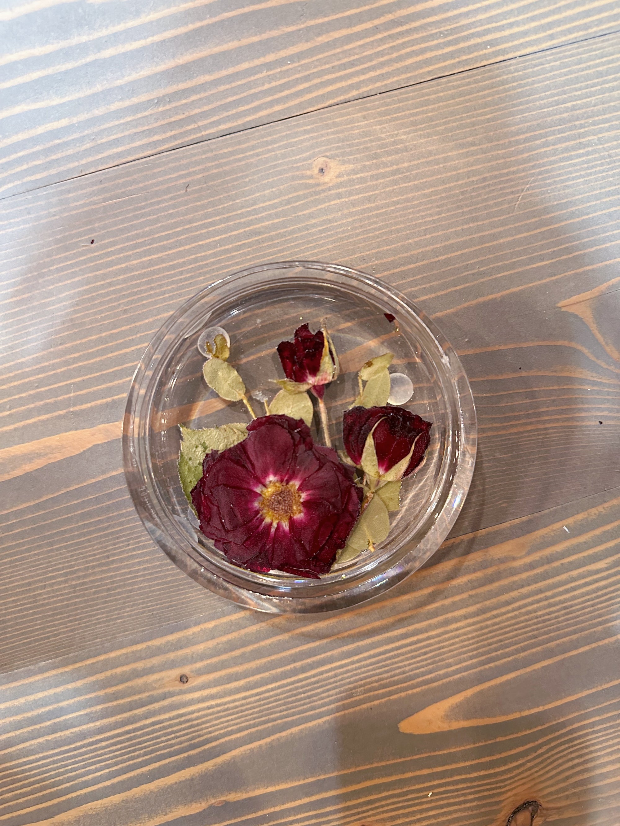 A circle resin ring dish with a pressed red rose, two spray roses, and a few green leaves is set on top of a wooden surface. 