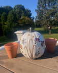 a white pumpkin with colorful pressed flowers on a table with other decorations outside with trees in the background