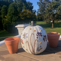 a white pumpkin with colorful pressed flowers on a table with other decorations outside with trees in the background