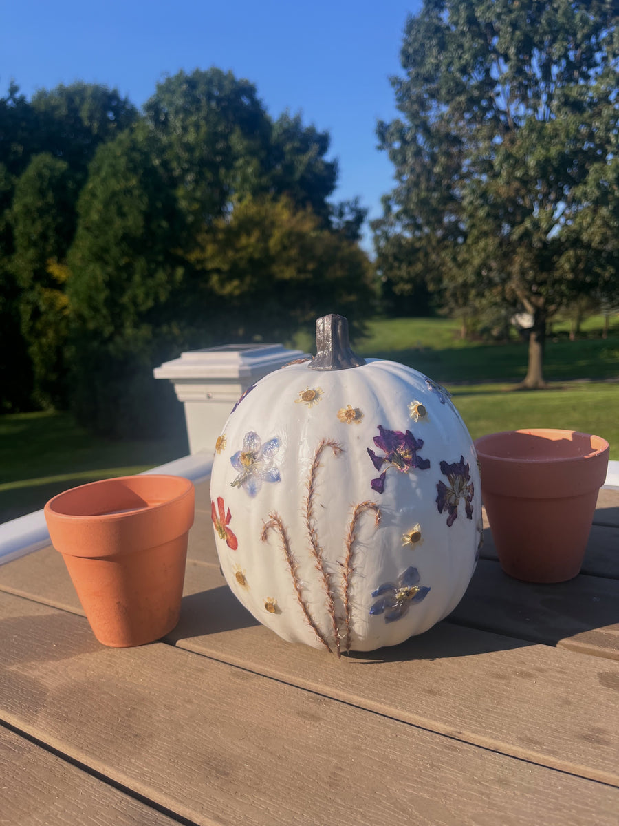 a white pumpkin with colorful pressed flowers on a table with other decorations outside with trees in the background