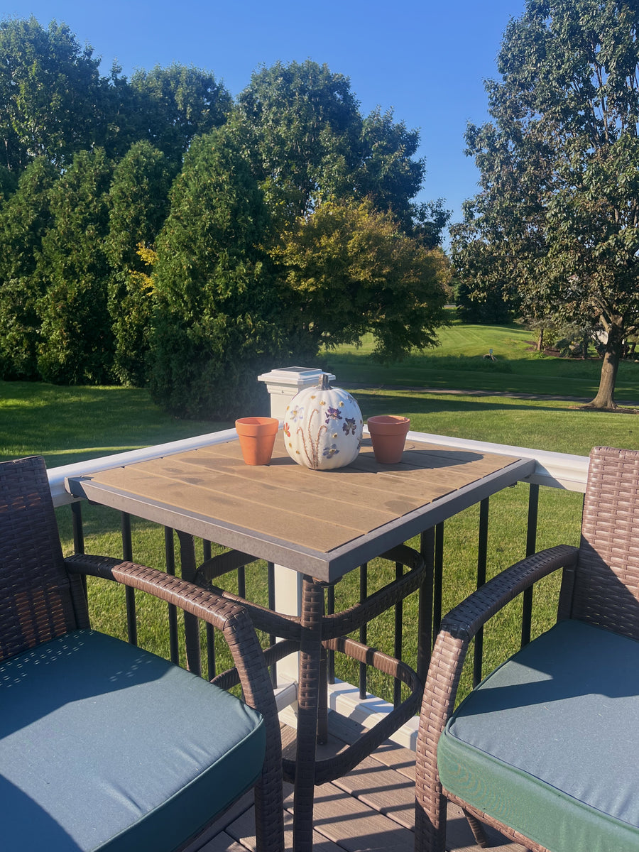 an outdoor porch with chairs and a table that has a white pumpkin with colorful pressed flowers on it and other decor with trees in the background