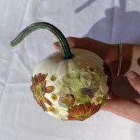 a small white pumpkin with pressed florals designed on it being held my someone on the table