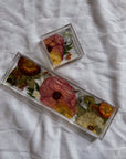 A square resin ring dish and a resin display tray with warm-colored pressed flowers and light greenery are placed on top of a white surface.