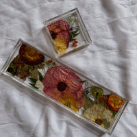 A square resin ring dish and a resin display tray with warm-colored pressed flowers and light greenery are placed on top of a white surface.