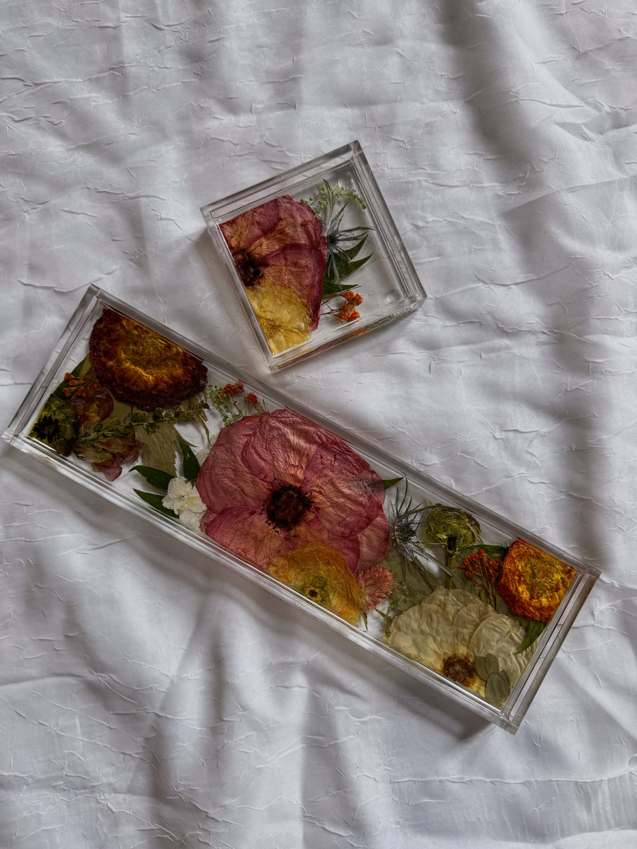 A square resin ring dish and a resin display tray with warm-colored pressed flowers and light greenery are placed on top of a white surface.