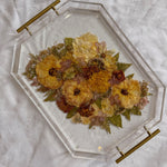 An autumn colored pressed bouquet is preserved in a geometric resin serving tray with gold handles, placed on top of a white surface. 