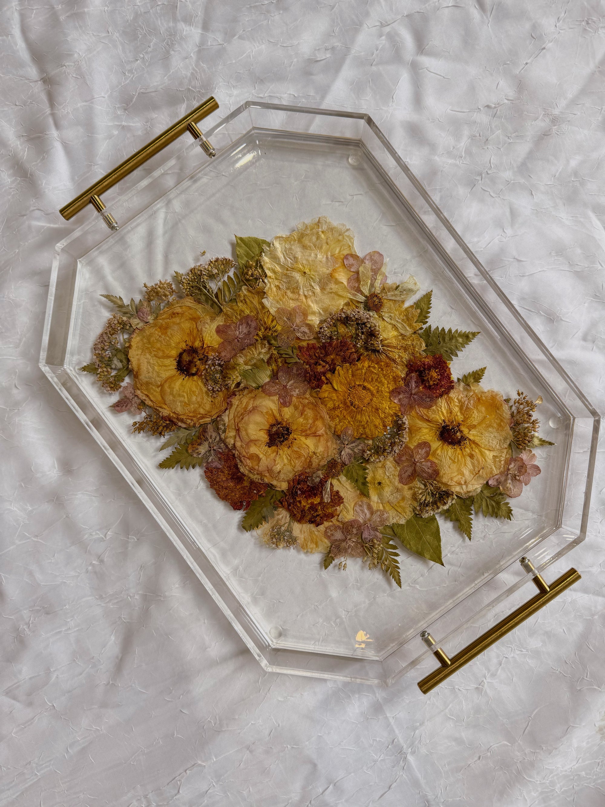 An autumn colored pressed bouquet is preserved in a geometric resin serving tray with gold handles, placed on top of a white surface. 