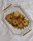 An autumn colored pressed bouquet is preserved in a geometric resin serving tray with gold handles, placed on top of a white surface. 