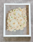 An all white ball bouquet pressed and preserved into a white frame against concrete.