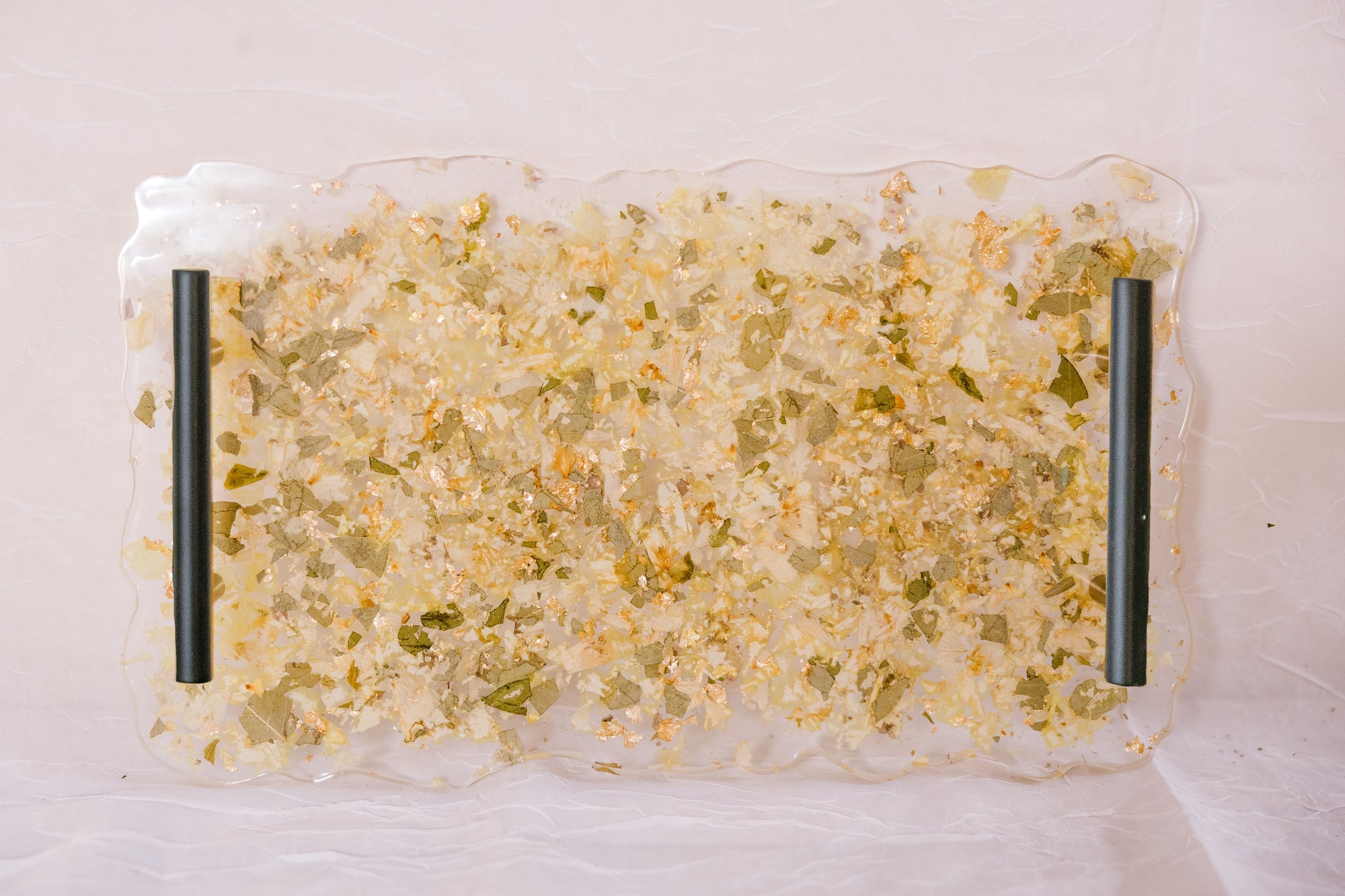 A resin serving tray with green leaves, white flowers, and gold metallic flakes are designed like confetti. The tray has black handles and is against a white background. 