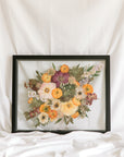 A colorful bouquet with purple, orange flowers with greenery are pressed inside this black wood floating frame.