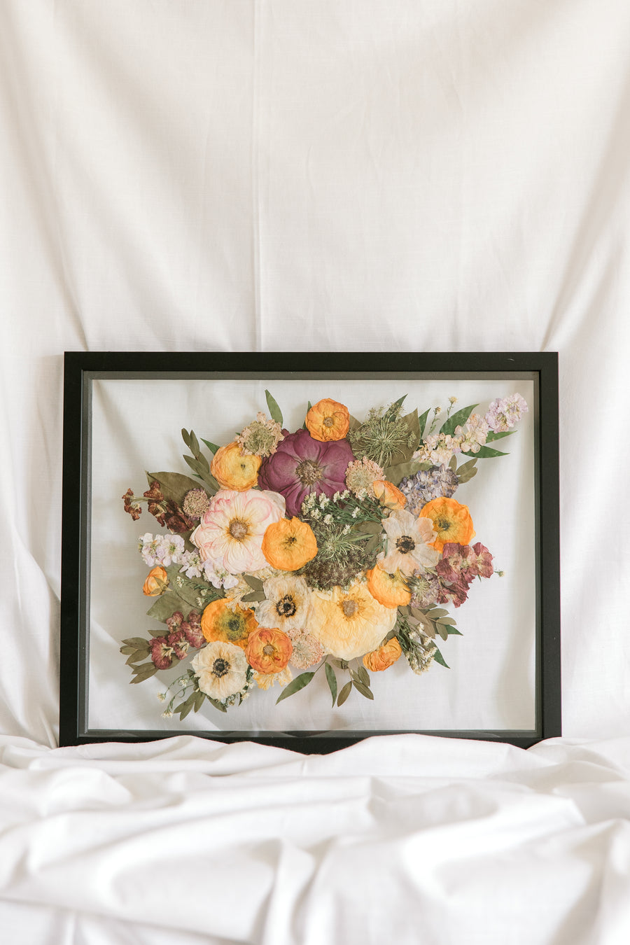 A colorful bouquet with purple, orange flowers with greenery are pressed inside this black wood floating frame.