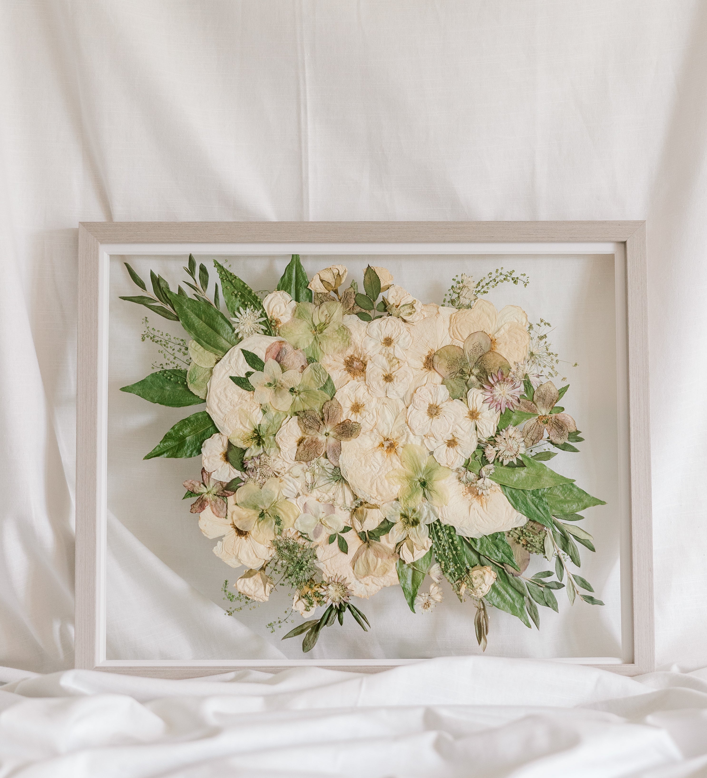 A white and green pressed wedding bouquet on display in a glass floating frame surrounded by gray wood.