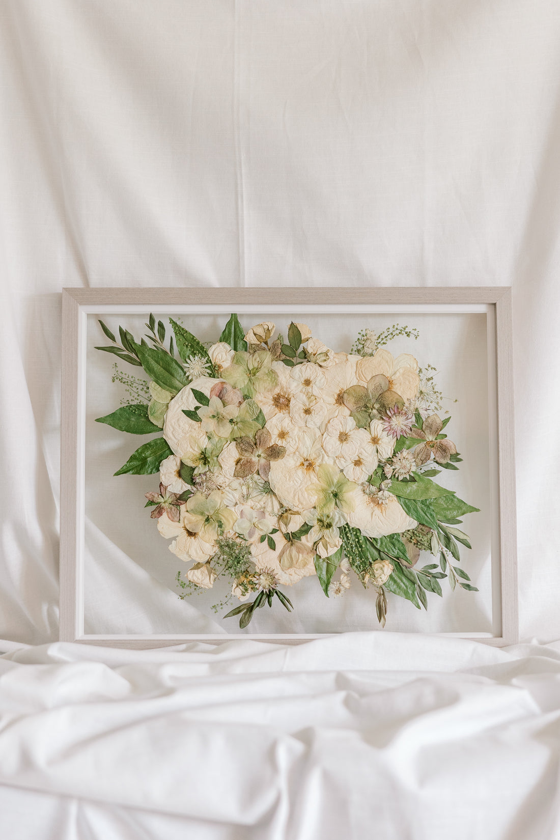 A white and green pressed wedding bouquet on display in a glass floating frame surrounded by gray wood.