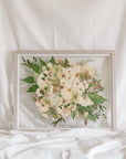 A white and green pressed wedding bouquet on display in a glass floating frame surrounded by gray wood.