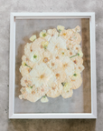 An all white ball bouquet pressed and preserved into a gray frame against concrete.