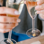 A couple is toasting two champagne glasses with real pressed flower drink charms around the stems above a wooden table. 