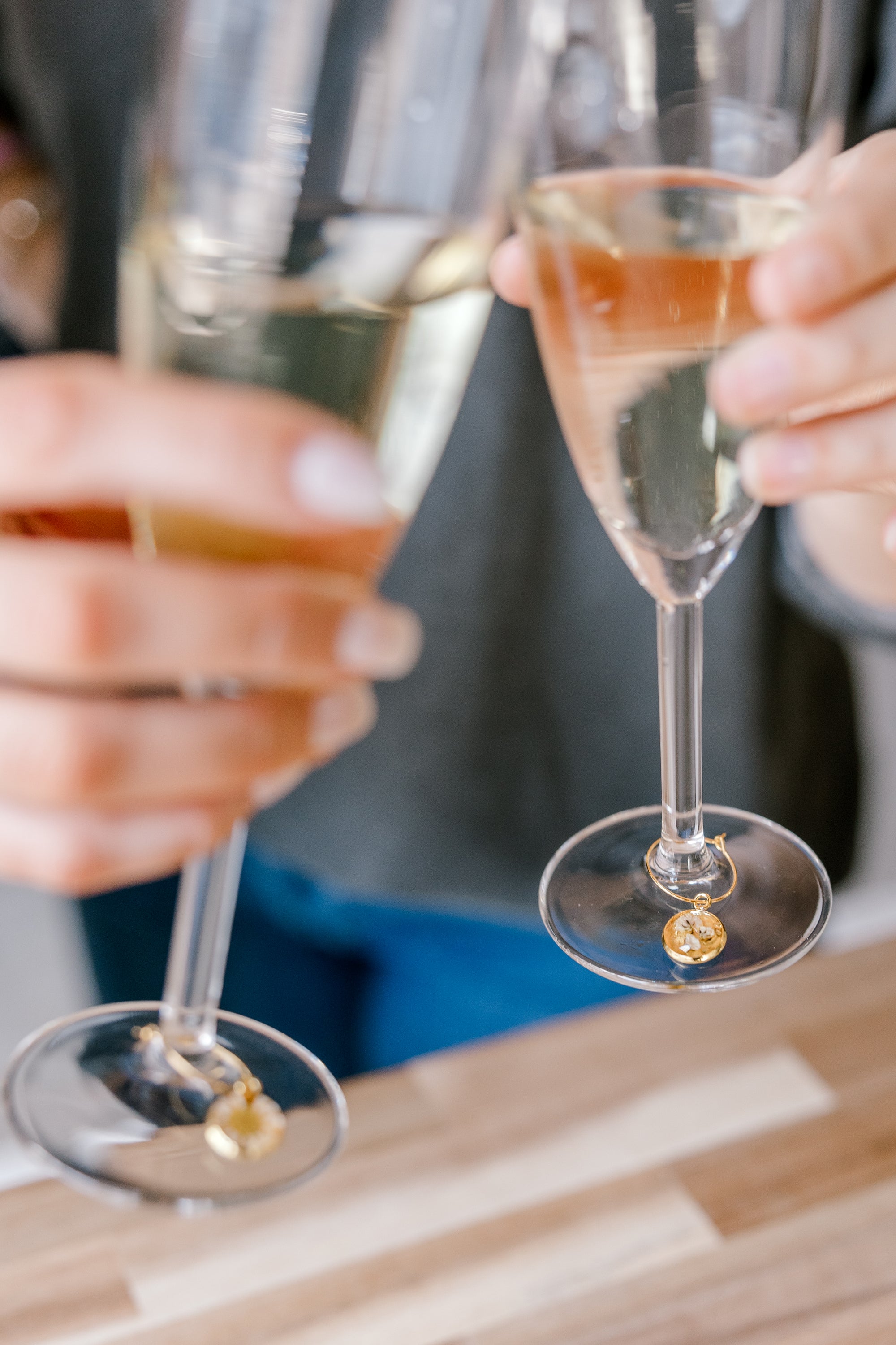 A couple is toasting two champagne glasses with real pressed flower drink charms around the stems above a wooden table. 