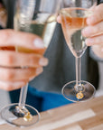 A couple is toasting two champagne glasses with real pressed flower drink charms around the stems above a wooden table. 