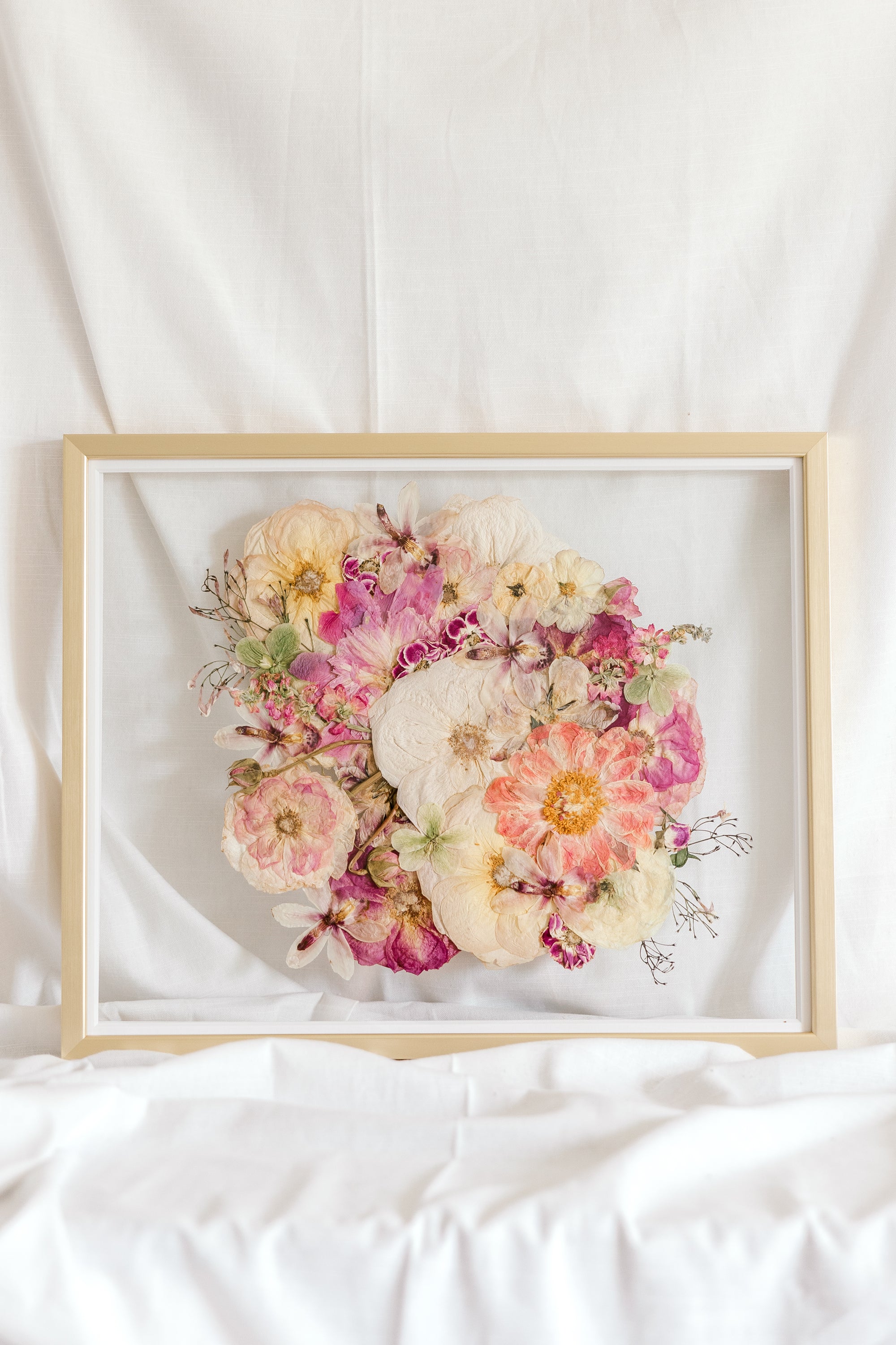 A bright pink and white pressed flower arrangement from a wedding bouquet is centered inside this gold wood frame.