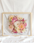 A bright pink and white pressed flower arrangement from a wedding bouquet is centered inside this gold wood frame.