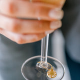 A woman is holding a glass of champagne with a pressed flower drink charm. The charm features a real small white flower. 