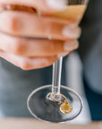 A woman is holding a glass of champagne with a pressed flower drink charm. The charm features a real small white flower. 