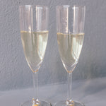 Two champagne glasses with pressed flower drink charms are placed next to each other against a grey background. The charms include real flowers. 